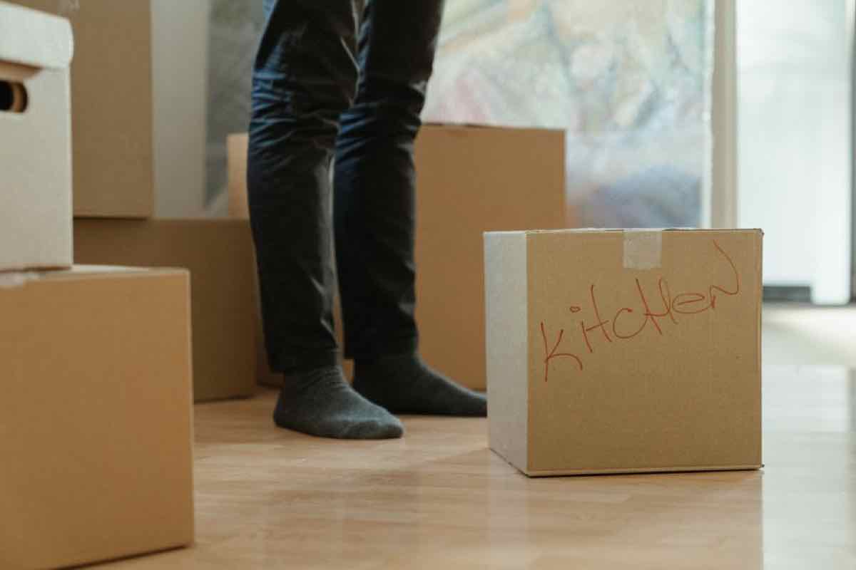 Cardboard moving box labeled 'kitchen' on a hardwood floor with a person's feet visible in the background