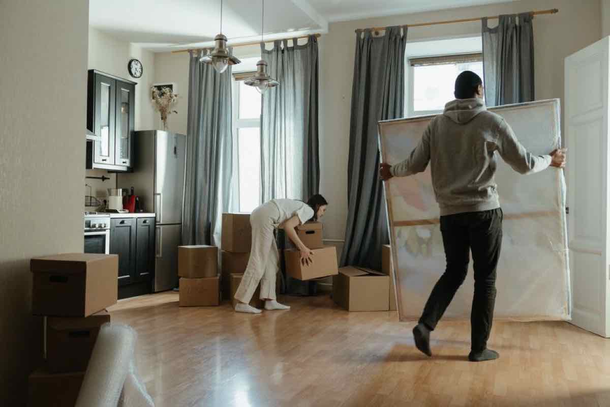Two people carrying moving boxes and a wrapped mirror through a living room